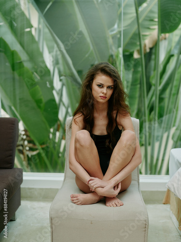 Young woman sitting on chair with knees drawn up, looking pensive and contemplative in a modern indoor setting