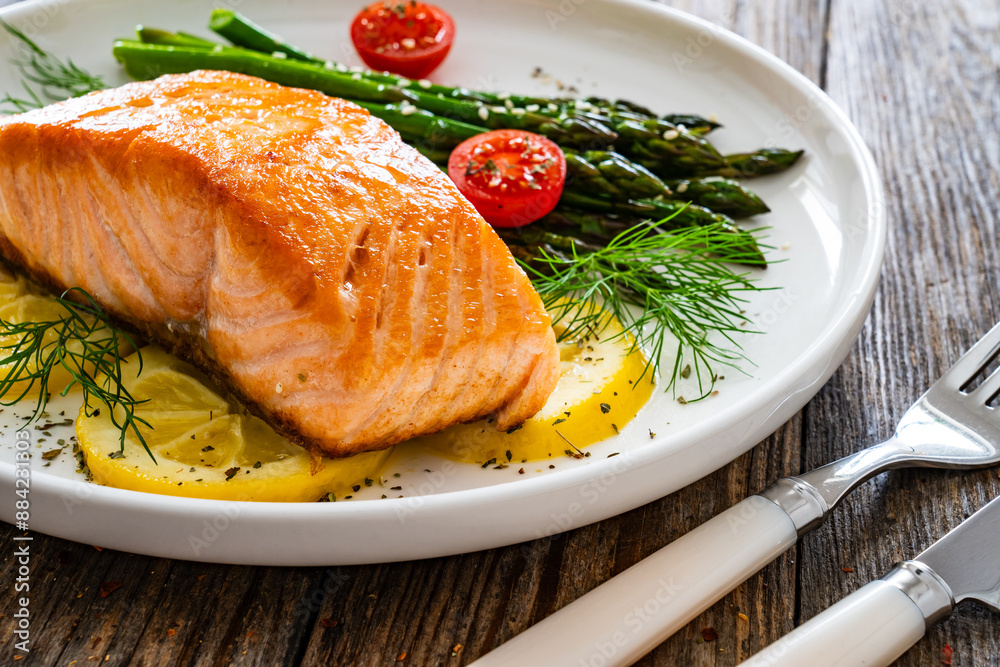 Fried salmon steak with cooked green asparagus, cherry tomatoes and lemon slices served on white plate on wooden table Fried salmon steak with cooked green asparagus, cherry tomatoes and lemon slices served on white plate on wooden table