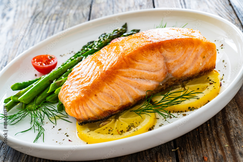 Fried salmon steak with cooked green asparagus, cherry tomatoes and lemon slices served on white plate on wooden table Fried salmon steak with cooked green asparagus, cherry tomatoes and lemon slices served on white plate on wooden table