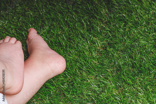  From above of anonymous kid with tender and delicate barefoot while sitting alone on green grass in park and in daylight 