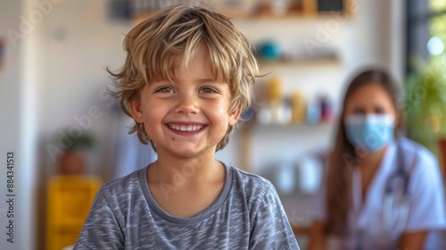 Smiling boy in casual attire enjoying an activity indoors with a caregiver in the background wearing a mask during a sunny day