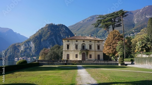 This image shows a grand villa situated by a tranquil lake surrounded by towering mountains under a crisp, blue sky. photo