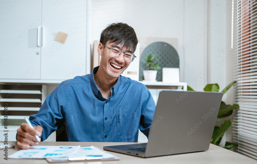Fototapeta premium emotional, notebook, freelance, job, learning, networking, news, worker, typing, working. A young man is sitting at a desk with a laptop and a stack of papers. He is smiling and he is happy.