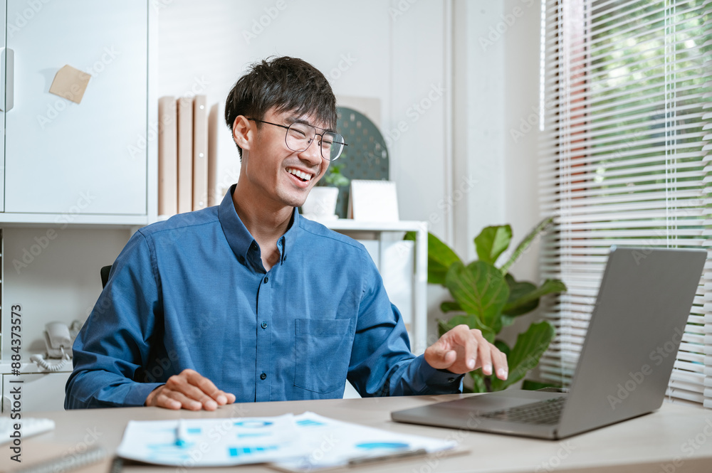 typing, analysis, document, executive, job, manager, paperwork, research, working, worker. A man is sitting at a desk with a laptop and smiling. He is wearing glasses and a blue shirt. typing, analysis, document, executive, job, manager, paperwork, research, working, worker. A man is sitting at a desk with a laptop and smiling. He is wearing glasses and a blue shirt.