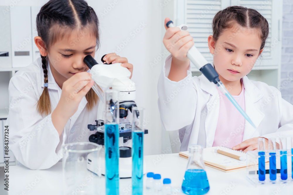 Two young girls, dressed in white lab coats, engage in scientific experiments in a laboratory. One girl examines samples through a microscope while the other uses a pipette to handle blue liquid Two young girls, dressed in white lab coats, engage in scientific experiments in a laboratory. One girl examines samples through a microscope while the other uses a pipette to handle blue liquid