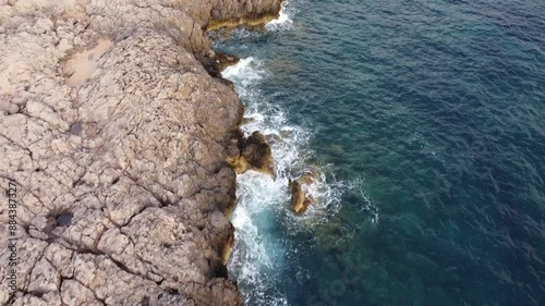 Aerial drone footage of a rocky coastline with waves crashing against the shore. Smooth top-down view of the rugged cliffs and deep blue sea. Perfect for travel, nature, and coastal themes.