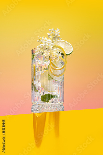 Homemade fresh elderflower lemonade garnished with lemon peel and a blooming branch on a bright pink and yellow background