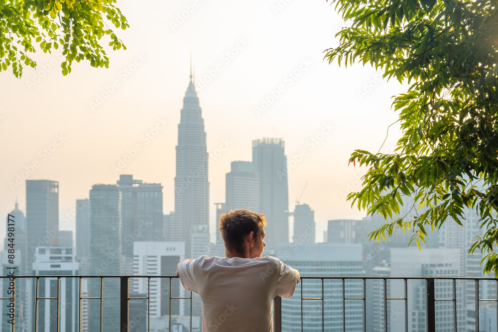 Fototapeta premium Back view of man tourist standing on the rooftop terrace of high rise building with green trees and looking at panoramic skyline of Kuala Lumpur city at sunrise, Malaysia. Rear view of male traveler