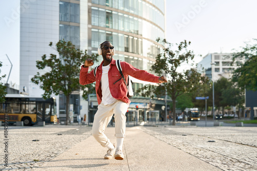 Happy cool young African Black man student hipster wearing headphones, sunglasses and backpack feeling joy having fun listening music and dancing on city street outdoors on sunny day, full body.