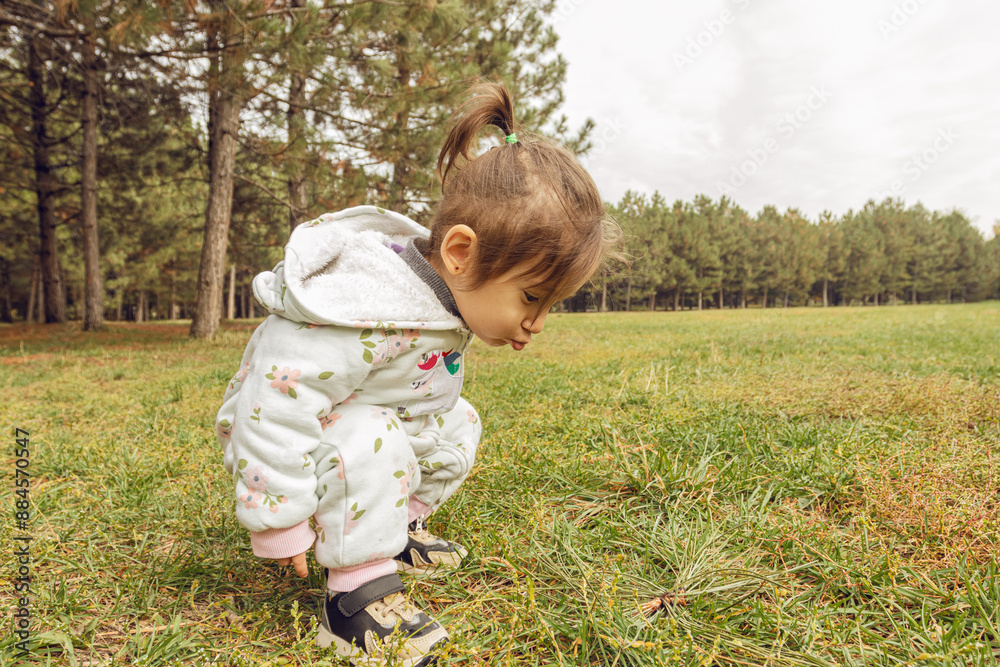 Fototapeta premium Cheerful toddler girl in white floral jumpsuit running in pine forest park. Family lifestyle, childhood development, or outdoor recreation themes.