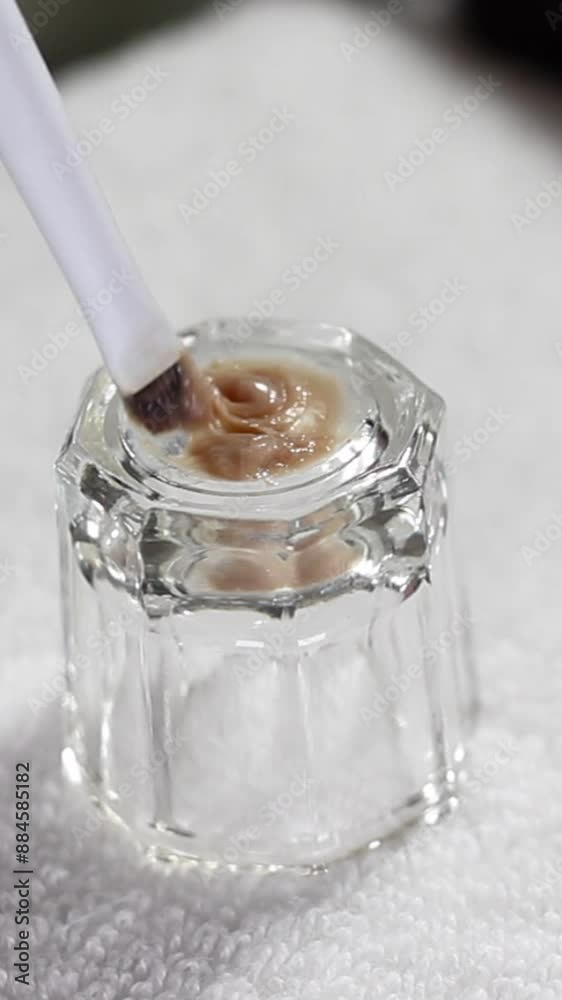 Close-up of a makeup brush being dipped into a small glass container with beige cosmetic product. The image highlights the preparation and application process in beauty treatments. Close-up of a makeup brush being dipped into a small glass container with beige cosmetic product. The image highlights the preparation and application process in beauty treatments.