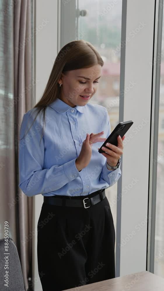 Business lady wearing blue shirt and black trousers standing at window in office. Woman writing a message on her smartphone. Blurred backdrop. Vertical video Business lady wearing blue shirt and black trousers standing at window in office. Woman writing a message on her smartphone. Blurred backdrop. Vertical video
