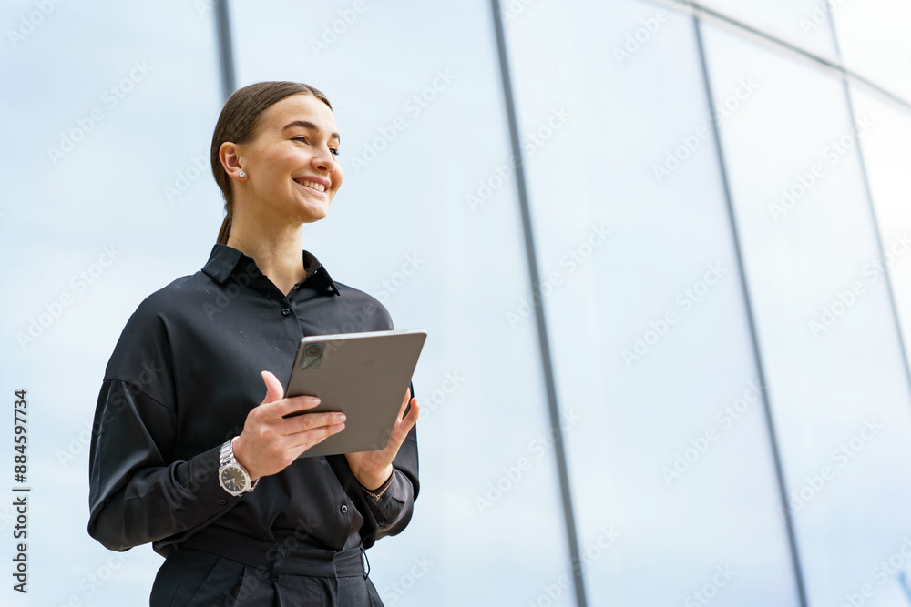 A young professional woman smiles while holding a tablet, standing against a reflective glass wall. A young professional woman smiles while holding a tablet, standing against a reflective glass wall.