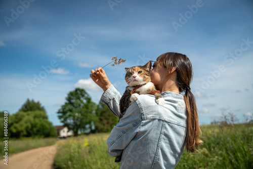 Smiling female owner hugging stern tricolor cat while walking through rural countryside under rays of hot summer sun. Back of girl wanting to play with plucked flower with tired uninterested kitten.