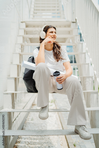 One young caucasian woman student sitting on stairs near university listening to music and drinking coffee 