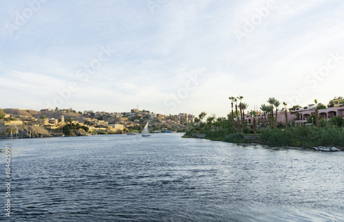 Aswan, Egypt - November 16, 2022: High angle and afternoon view of a sailboat sailing on Nile River against downtown houses and palm trees 