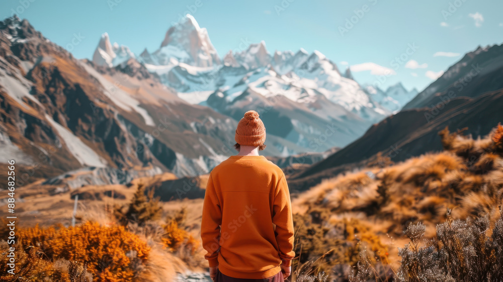 A photograph of a person in an orange sweater and beanie, facing away from the camera, standing on a mountain trail, with snow-capped peaks in the background A photograph of a person in an orange sweater and beanie, facing away from the camera, standing on a mountain trail, with snow-capped peaks in the background
