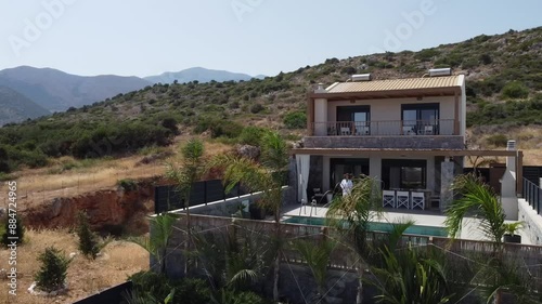 Dark haired slim woman walks to a private pool and takes off the bathrobe and sits down in a swimming costume to the pool - villa in Greece Crete and palm trees in the foreground can be seen photo