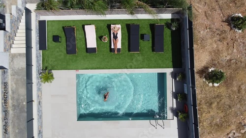 Drone flies over an outdoor pool in bird's eye view - Woman in swimming costume lies on sunbed and man jumps into the pool - Villa in Greece Crete with palm trees photo