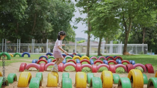 Asian young girl is playing in a park with a variety of playground equipment. She is standing on a tire and she is having fun. The park is filled with colorful and interesting play equipment
