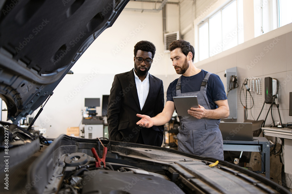 Mechanic in work overalls explaining car issues to customer in suit in modern auto repair shop. Professional consultation about vehicle maintenance and repair process. Mechanic in work overalls explaining car issues to customer in suit in modern auto repair shop. Professional consultation about vehicle maintenance and repair process.
