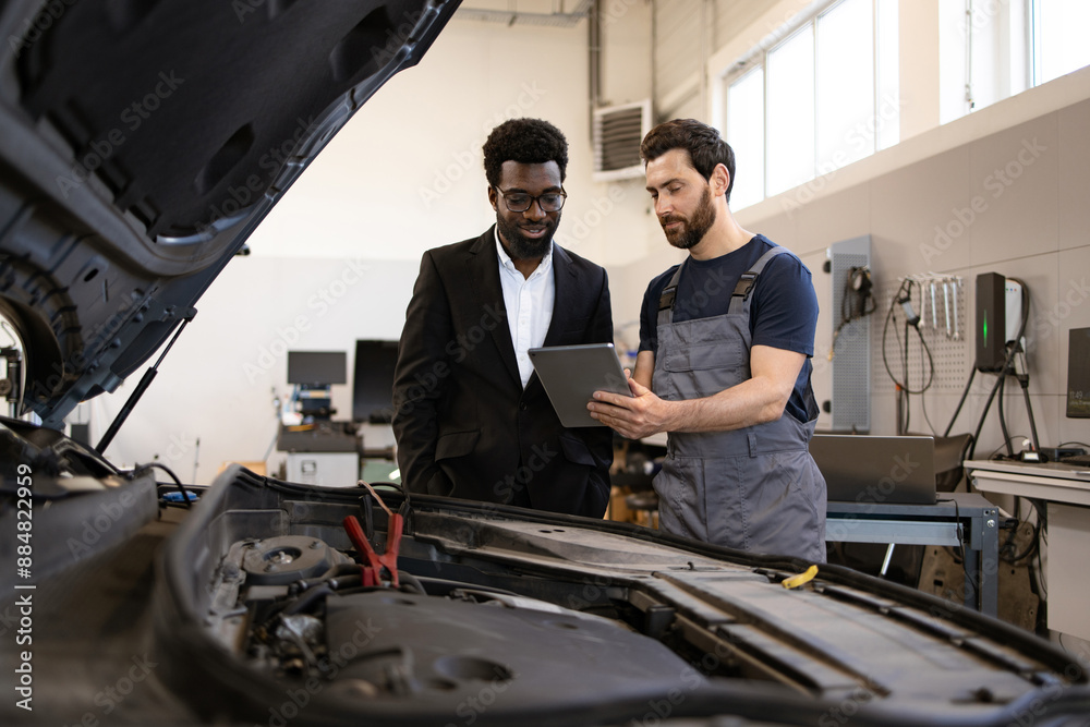 Mechanic in overalls discussing car maintenance with businessman using tablet. Scene in modern auto repair shop with open car hood. Concept of professional automotive service and customer service. Mechanic in overalls discussing car maintenance with businessman using tablet. Scene in modern auto repair shop with open car hood. Concept of professional automotive service and customer service.