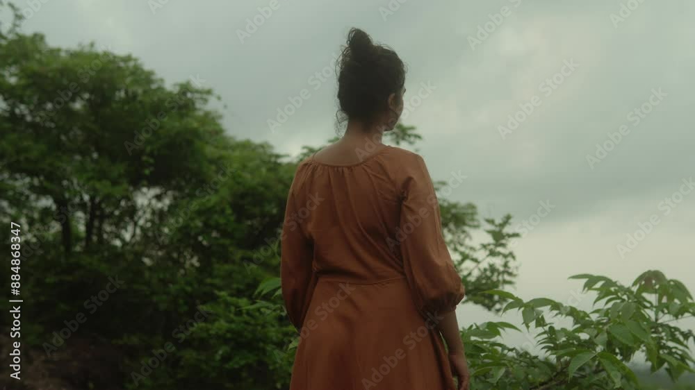Woman in an orange dress stands in a lush green forest under a cloudy sky Woman in an orange dress stands in a lush green forest under a cloudy sky