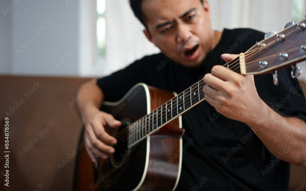 Obraz premium Asian man with sitting on a sofa in his home's living room, picking up a guitar.