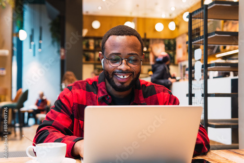 Candid shot of attractive African American student in checkered shirt sitting at cafe table with mug, keyboarding on laptop pc, typing comments online while browsing social networks