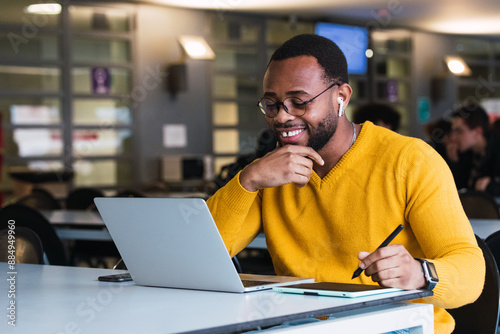 Indoor office shot of cheerful happy young African American man with yellow sweater sitting in front of open laptop wearing earphones while having video conference call with business partners