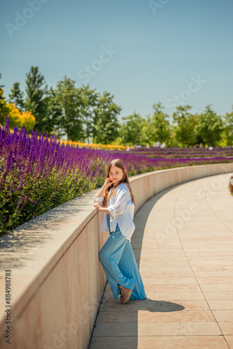 A young girl is basking in the warm rays of the sun, enjoying the beauty of the park, overgrown with bright purple sage. Her white shirt and blue trousers complete the landscape