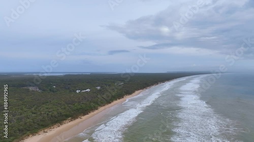 Flight heads north up Rainbow Beach with holiday homes surrounded in lush woodlands as light hazy mist approaches from K'gari Fraser Island. Pulsating subdued green breakers crash onto the coast. photo