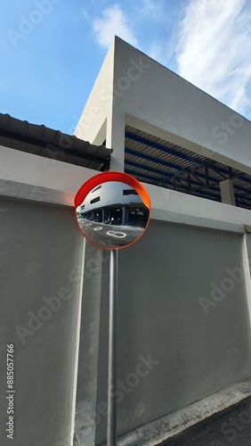 a convex safety mirror mounted on a pole, reflecting a parking area with motorcycles. The mirror is attached to a gray wall of a modern building under a blue sky with some clouds.