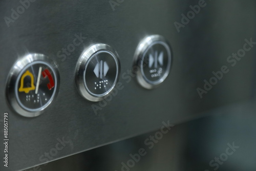 a close-up of a metallic surface with three circular buttons. Each button features different icons: an elevator emergency button, open button, and close button.