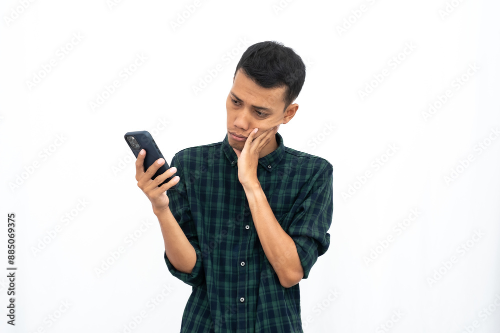 Asian man wearing a casual green checkered shirt is stressed or dizzy from getting bad news when looking at the smartphone screen. Isolated white background. Asian man wearing a casual green checkered shirt is stressed or dizzy from getting bad news when looking at the smartphone screen. Isolated white background.