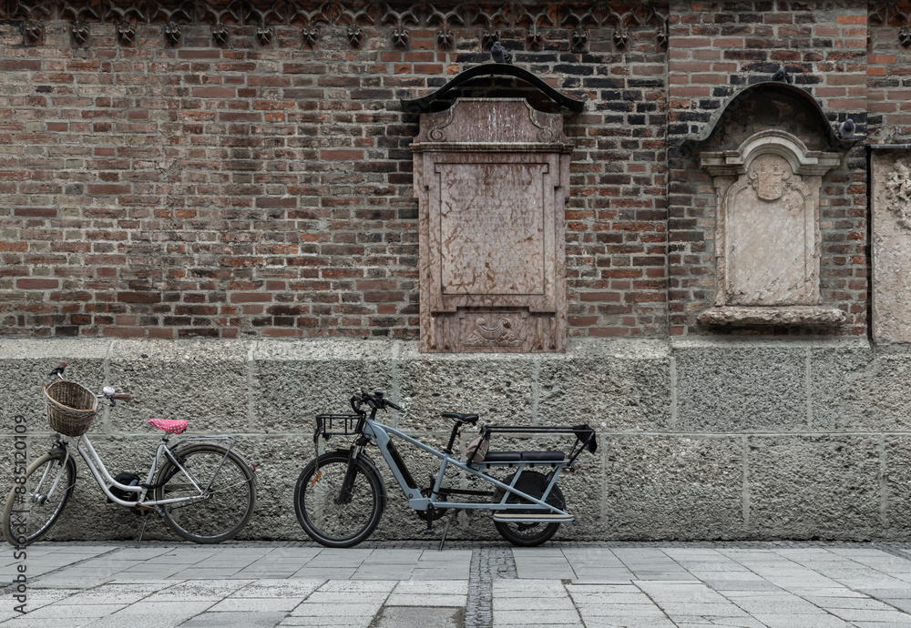 Vintage bike with basket and Electric longtail cargo bike are parked side of Mounted burial headstones based upon the outside Old brick wall of the Munich Frauenkirche or Frauenplatz. Cathedral Church