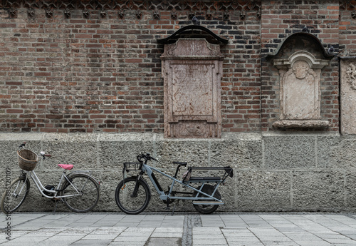 Obraz na płótnie Vintage bike with basket and Electric longtail cargo bike are parked side of Mounted burial headstones based upon the outside Old brick wall of the Munich Frauenkirche or Frauenplatz. Cathedral Church w salonie