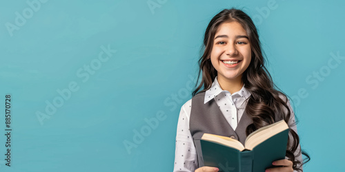 A cheerful teenage girl in a white shirt and grey vest happily smiles, holding an open book against a blue background with enthusiasm and joy