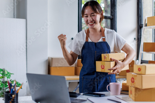 Happy young asian woman startup small business freelance holding parcel box and computer laptop and sitting on chair, Online marketing packing SME box delivery concept