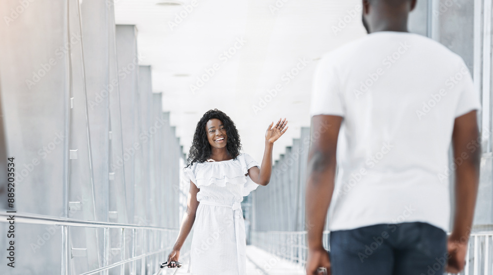 Black woman in a white dress waves goodbye to a man in a white shirt in an airport terminal. The man is turned away from the camera, and the woman is looking directly at the camera with a smile.