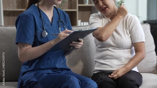 Young Asian nurse attends to older grey-haired mature woman sitting on sofa, conducting health assessment and discussing conditions osteoporosis, arthritis, Alzheimer's disease, dementia, and more.