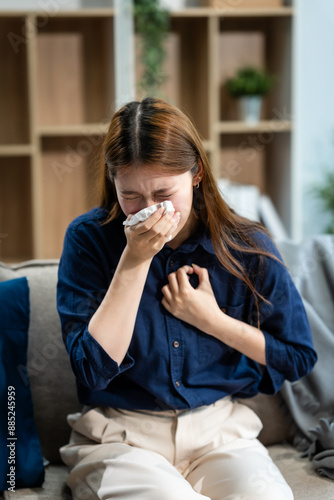 A young Asian woman sits on a sofa, feeling sick with a cold. Wrapped in a blanket, she has the flu, coughs, and uses tissue paper for her runny nose at home. immune system