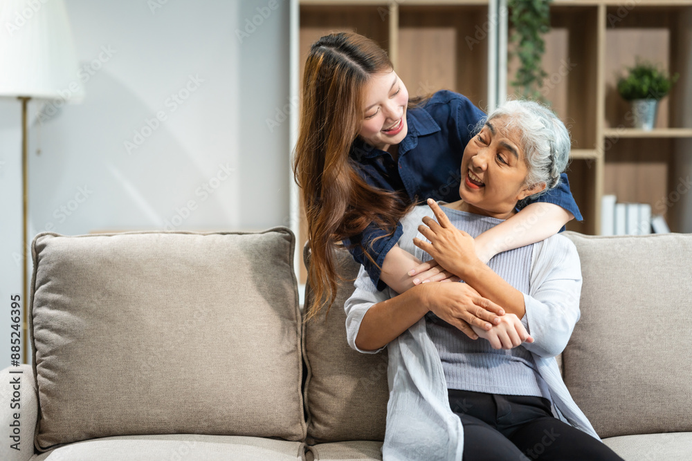 A mature mom and a young Asian woman, mother and daughter, sit together on a sofa in their living room, celebrating Mother's Day with love and bonding at home. A mature mom and a young Asian woman, mother and daughter, sit together on a sofa in their living room, celebrating Mother's Day with love and bonding at home.