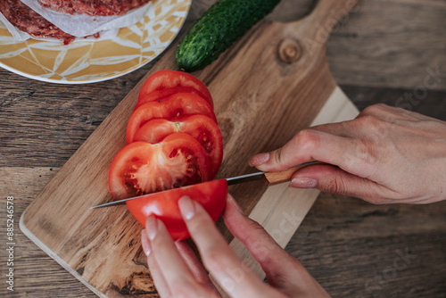 Hands of woman slicing tomato with knife on cutting board in kitchen photo