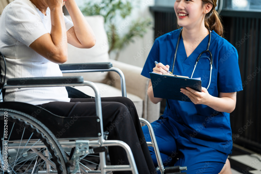 A young Asian nurse, the only woman present, assists an older grey-haired mature woman on a wheelchair sitting on a sofa. They focus on taking a medical history and conducting health checks. A young Asian nurse, the only woman present, assists an older grey-haired mature woman on a wheelchair sitting on a sofa. They focus on taking a medical history and conducting health checks.