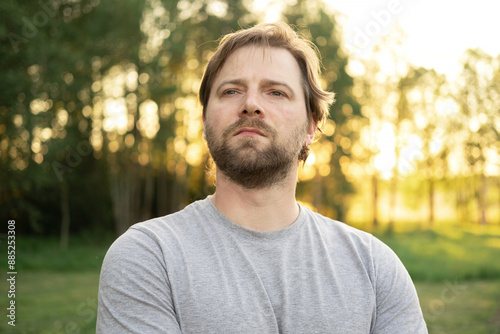 Portrait of handsome young man in gray t-shirt outdoors. Male with long hair and beard feel happy, calm and serious on Green nature sunset background. Man looking away into distance. Diversity People
