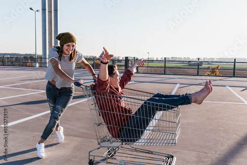 Young woman pushing boyfriend sitting in shopping cart at parking lot photo