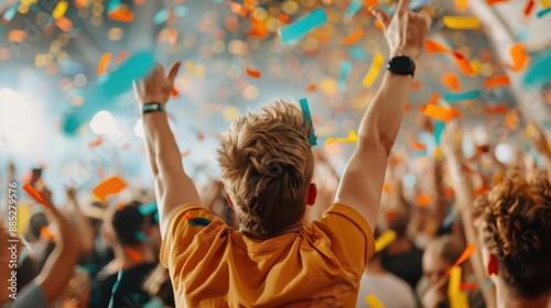 A man with blonde hair raises his arms in celebration amidst a crowd at a lively concert, with colorful confetti falling all around, capturing the joy and excitement of the moment. photo