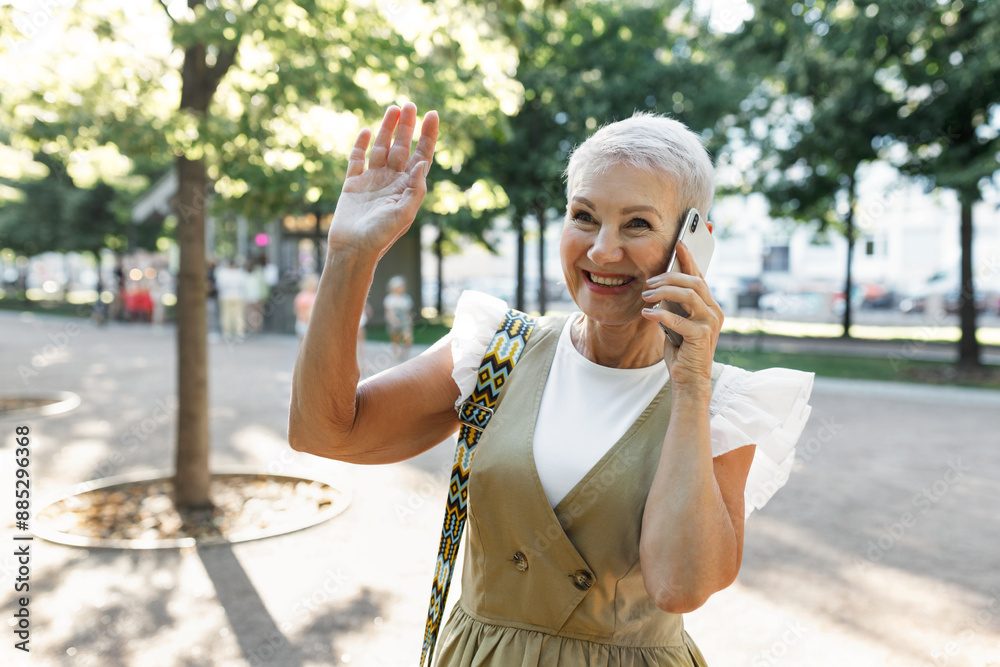 Obraz premium Outdoor portrait of happy pretty elegant caucasian female of 60s in elegant dress calling to her friend to find her in public park and greeting her bestie waving hand, walking on hot summer day