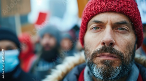 A serious-looking man, wearing a red knit cap and a warm jacket, stands amidst a crowd, likely at a protest or rally, conveying determination and resolve.